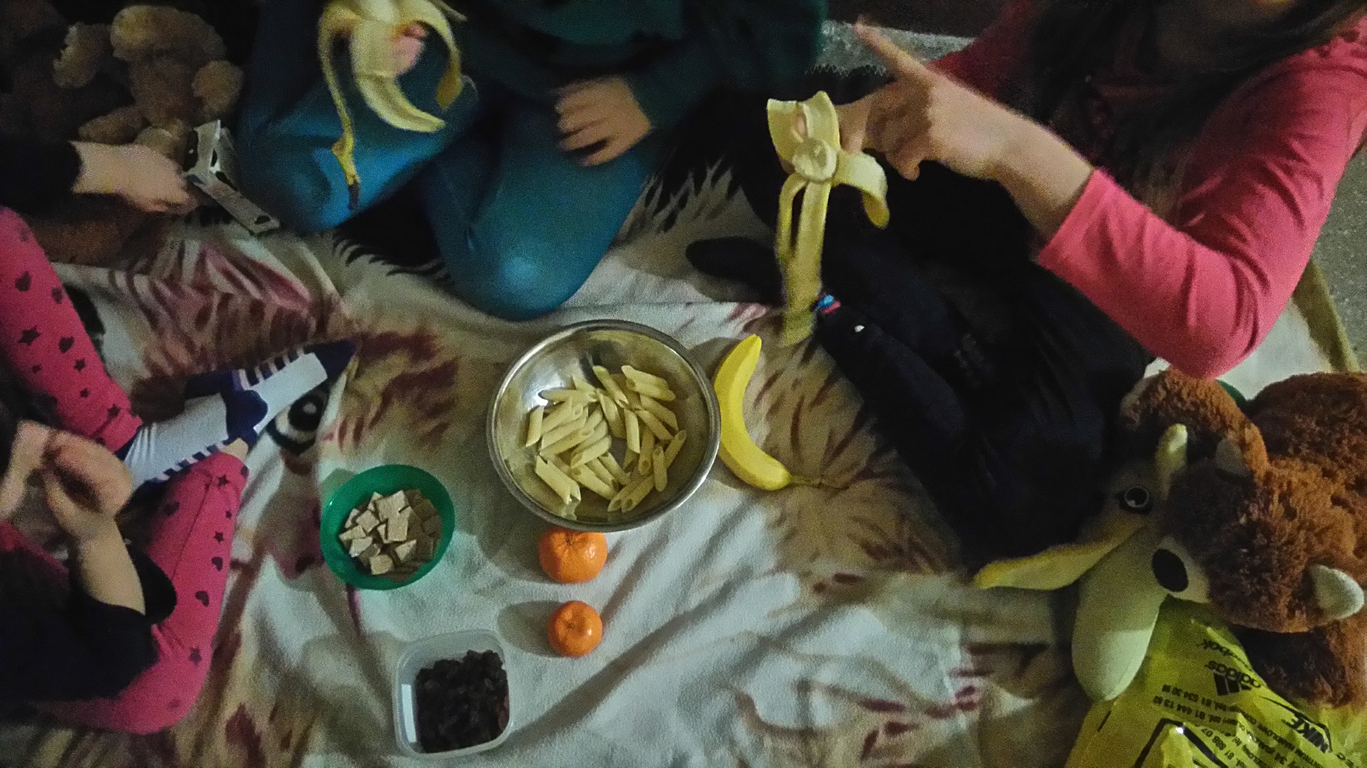 makaron, banany, mandarynki, płatki kukurydziane w zielonej misce leżą na piknikowym kocu, dzieci jedzące banany
