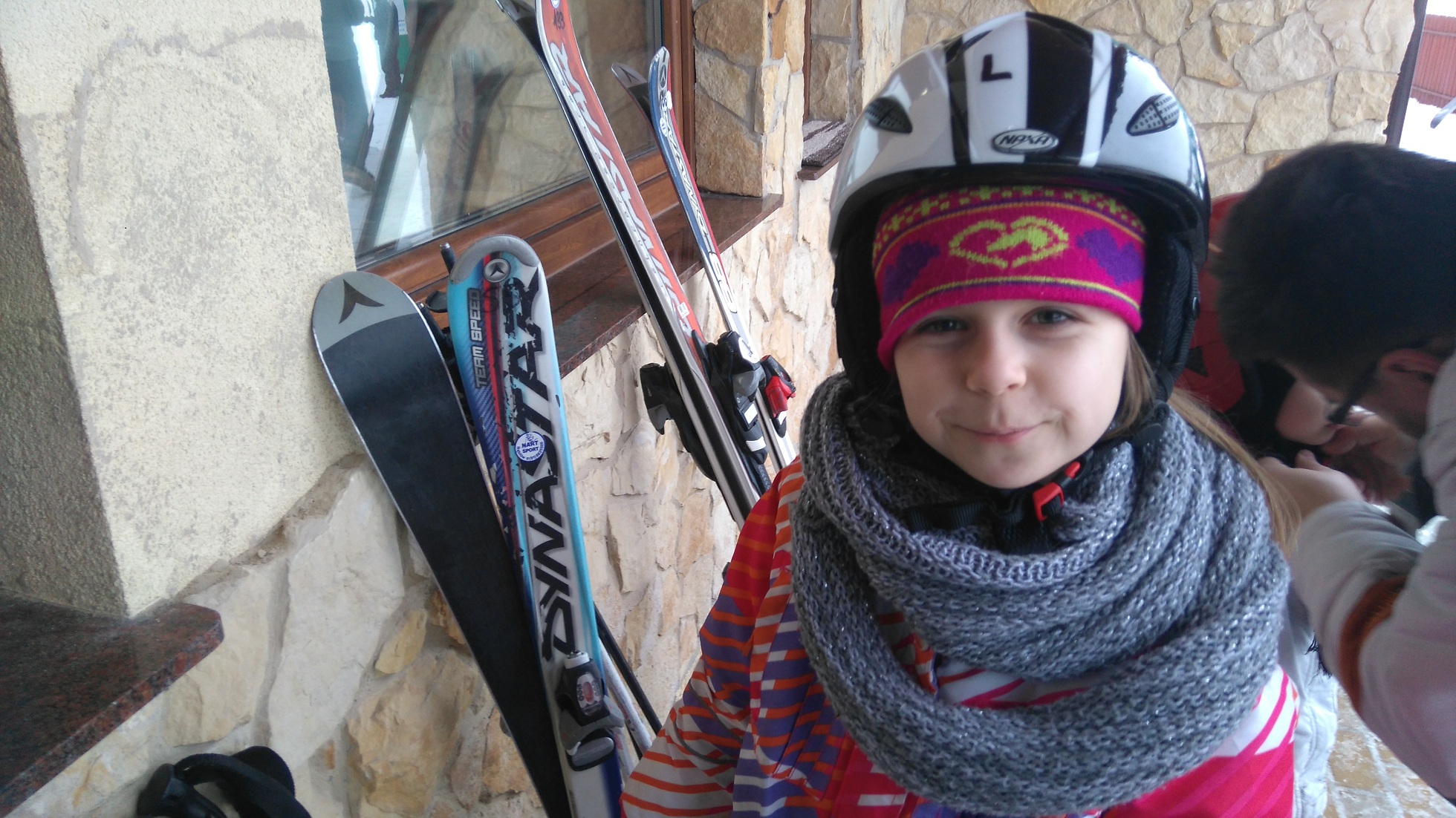 dziewczynka w stroju narciarskim, obok narty ooarte o ścianę budynku
